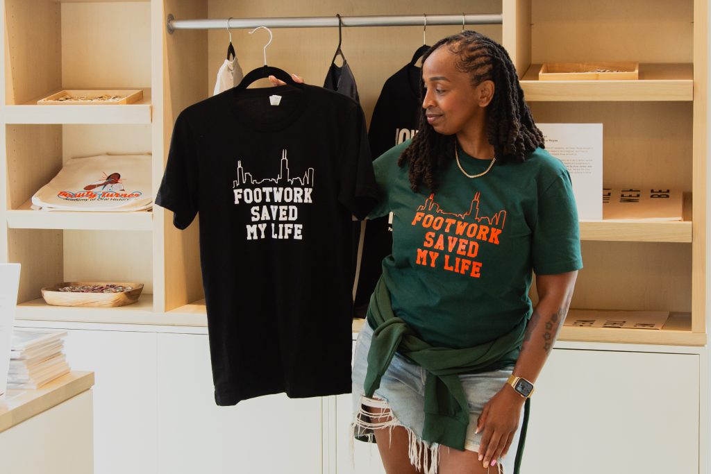 Dr. ShaDawn Battle stands in the gift shop of the National Public Housing Museum holding up a black "Footwork Saved my Life" shirt with the Chicago skyline illustrated with a single line. She is smiling while looking at it. She is wearing a green version of the shirt she is holds. Image by Joshua Clay Johnson.