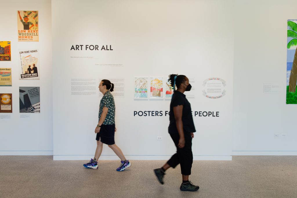 Liú méi zhì huì and jellystone robinson frazier walk in opposite direction in front the of the "Art for All, Posters for the People" exhibit at the National Public Housing Museum. Liú méi zhì huì is dressed in a button down green shirt with white polka dots, black bottoms, and colorful sneakers. jellystone robinson frazier is wearing a black button down, black capris, and black sneakers. Image by Joshua Clay Johnson.