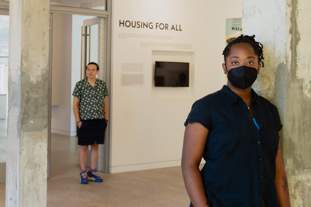 Liú méi zhì huì (pictured left) wears a green shirt with a white and yellow plant pattern, black shorts, colorful sneakers, and a ponytail. Liú stands in a doorway of an exhibition space in the National Public Housing Museum next to a section of the exhibition titled, "Housing for All," which is spelled out in black lettering above exhibition informational text and a built-in tv screen on the wall under the lettering. jellystone robinson frazier (pictured right) wears a black shirt with a blue undershirt, a black n95 mask and braids wrapped up in a ponytail. jellystone stands on a white and gray cement support beam holding up the gallery. The floors of the gallery are light, brown wood. Image by Joshua Clay Johnson.