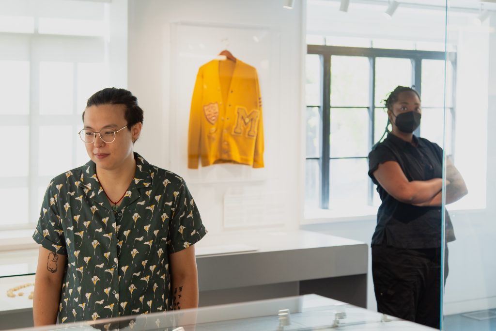 Liú méi zhì huì (pictured left) and jellystone robinson frazier (pictured right) pose in an exhibition space at the National Public Housing Museum. Liú méi zhì huì wears gold half-rimmed glasses, a green shirt with a white plant motif pattern, while jellystone robinson frazier wears a black short-sleeved blouse, capris, and an N95 mask. Between them a yellow letterman jacket hangs on view in the exhibition space. Behind them are large windows. Image by Joshua Clay Johnson.