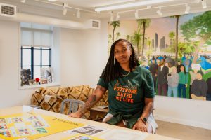 Dr. ShaDawn Battle wearing a green shirt with orange lettering of the words, "Footwork Saved My Life" sits on a chair at a wood desk inside the National Public Housing Museum. Behind Dr. Battle is a painting of people from diverse backgrounds standing in front of palm trees and a silver cityscape. Image by Joshua Clay Johnson.