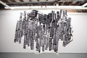 江麟冶《Scavenger Hunt》展览现场一景。展厅中央悬挂的是雕塑作品《摘录自父亲小说〈深圳故事〉》（2025），由父亲书写的文字片段构成，并封入树脂之中。文字选自父亲匿名发表于中国网络论坛的男同性恋小说《深圳故事》，连载时间为 2012 至 2017 年，直到论坛关闭。这些文字被印在相纸上，使用黑体——一种让艺术家想起青春年代的字体。它们已不再是完整的句子，而是留存在记忆中的片段，陪伴她一路寻迹。照片由艺术家提供。