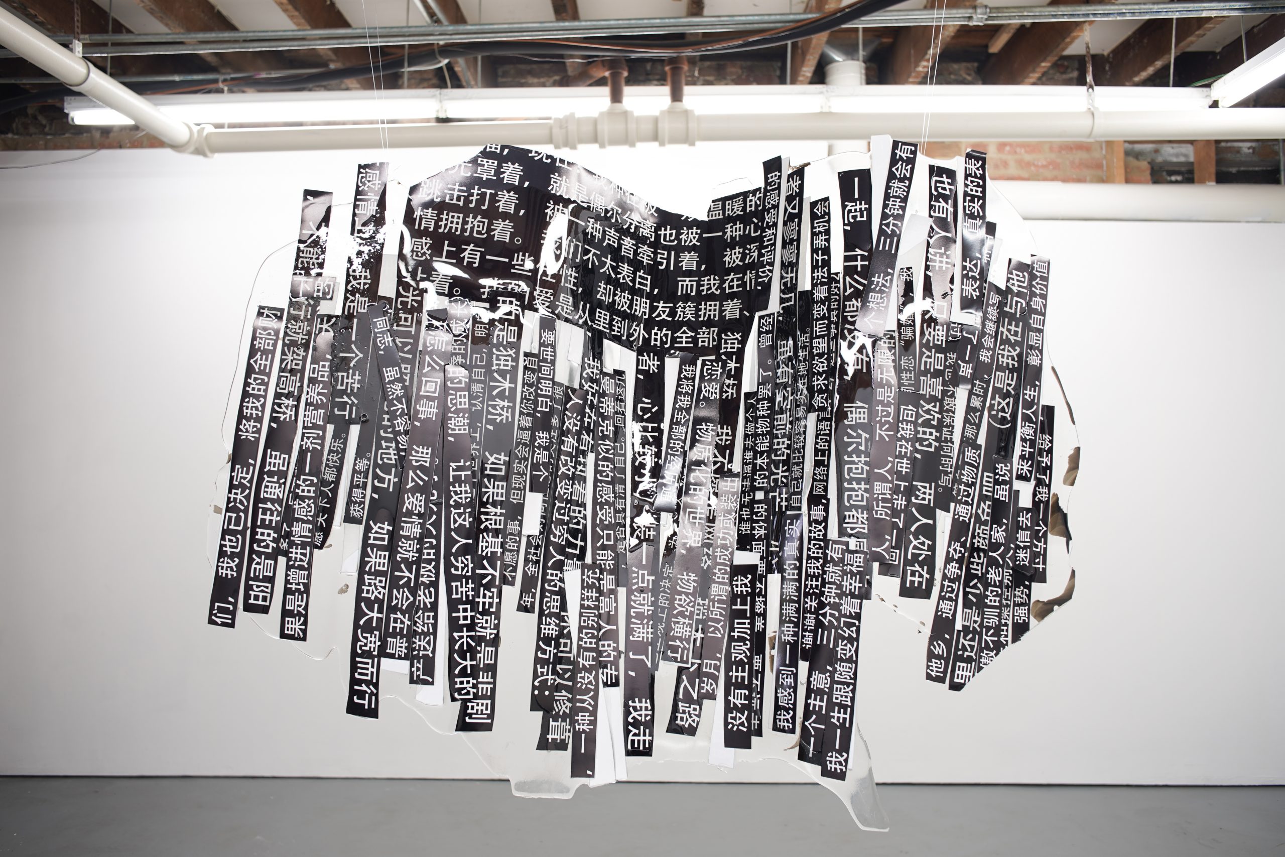 江麟冶《Scavenger Hunt》展览现场一景。展厅中央悬挂的是雕塑作品《摘录自父亲小说〈深圳故事〉》（2025），由父亲书写的文字片段构成，并封入树脂之中。文字选自父亲匿名发表于中国网络论坛的男同性恋小说《深圳故事》，连载时间为 2012 至 2017 年，直到论坛关闭。这些文字被印在相纸上，使用黑体——一种让艺术家想起青春年代的字体。它们已不再是完整的句子，而是留存在记忆中的片段，陪伴她一路寻迹。照片由艺术家提供。