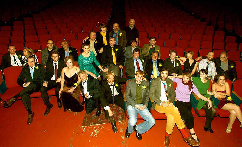A brightly lit image of an ensemble of 20 or so well dressed people of various ages. The group is seated in the first few rows of an empty theater smiling up toward the stage. the stage. The author Maggie is at the far right.