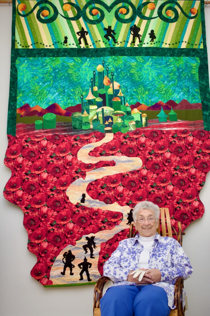 Portrait of Grace Stinton sitting in a chair in front of one of her large-scale picturesque quilts. The quilt features a scene from the Wizard of Oz as the protagonists go toward Emerald City. Image courtesy of Veronica Kirin.