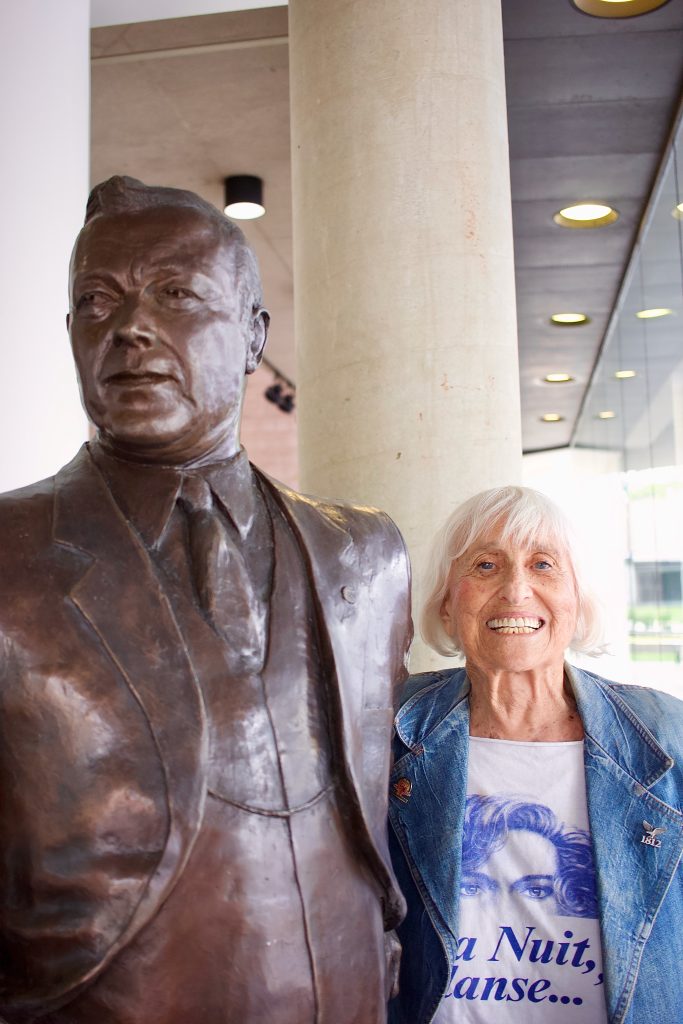 Portrait of Harriet Berg smiling brightly at the camera as she stands next to a bronze statue of a man in a three-piece suit. Image courtesy of Veronica Kirin.