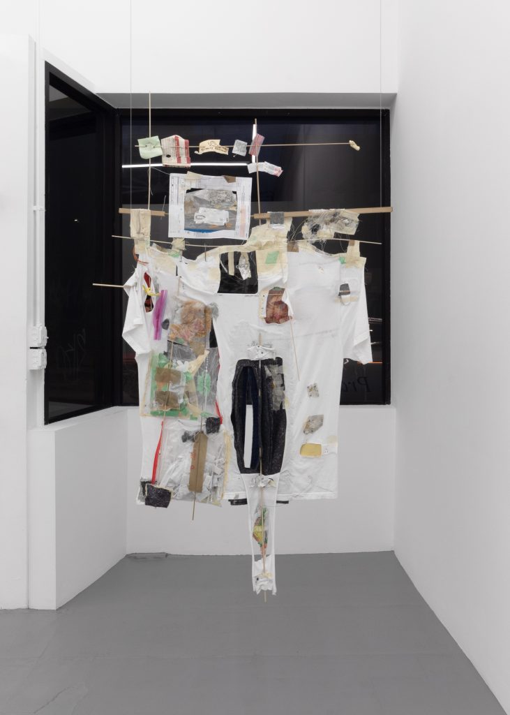 A white cotton t-shirt is suspended from the ceiling on a wooden dowel rod frame in front of a window. The t-shirt is interwoven with various materials, including a medical bill, gingko leaves, a dirty government document, newsprint, an alcohol swab with a blood stain, alcohol swab wrappers, seeds, fiberglass insulation, syringes and their wrappers, wood dust, fishing line, a kid’s jacket remnant with a metal zipper, thread, tape, quilting pins, etc.