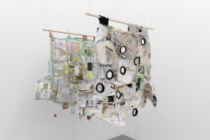 Two cotton t-shirts are suspended upside down from wooden dowel rods and interwoven with various materials, including tape, silicon caulk, thread, quilting pins, printer paper, various fabrics, plastic bags, etc. The t-shirt closer to the camera is punctured by a number of silicon rubber plumbing gaskets.