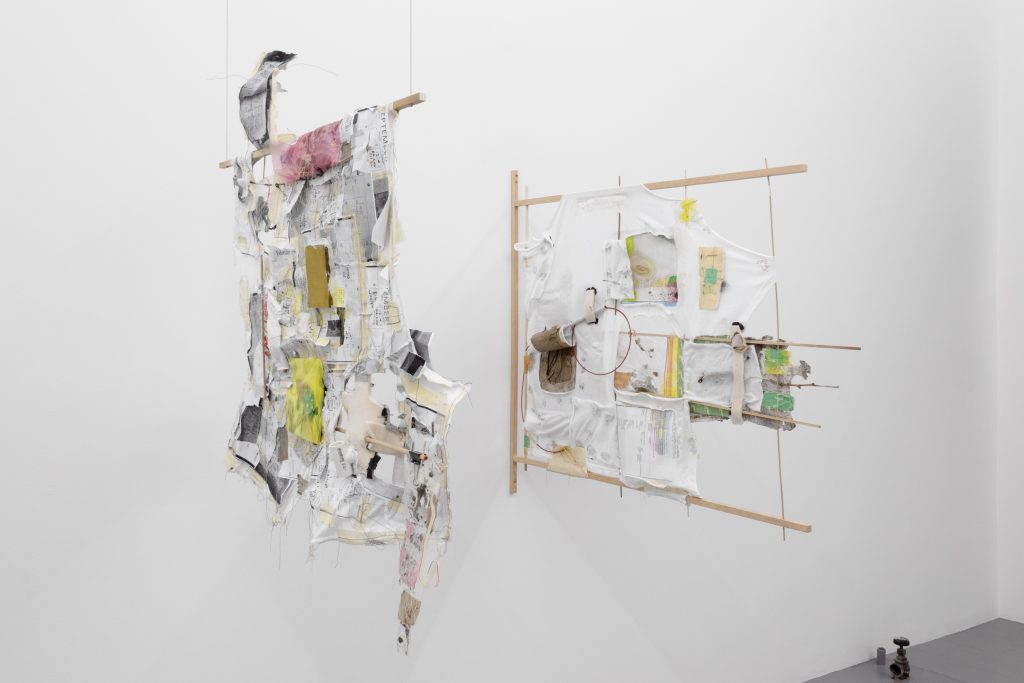 Two white cotton t-shirts are stretched over wooden dowel rod structures attached to the wall and suspended from the ceiling. The t-shirts are interwoven with various materials,
including tape, silicon caulk, thread, quilting pins, Ziploc bags, run-over cardboard boxes, eroded journal pages, alcohol swabs, seeds, cicada shells, Ricola wrappers, sanding sponge, binder clips, etc.