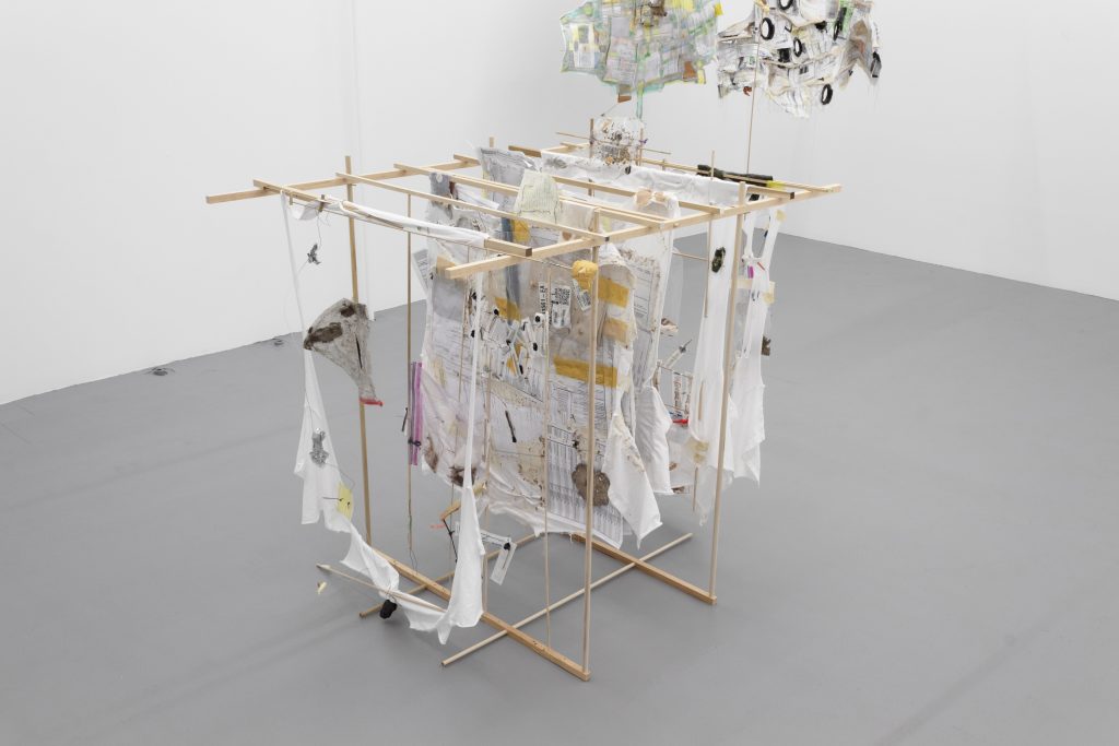 Half a dozen white cotton t-shirts are suspended from a wooden dowel rod structure resembling a clothes drying rack. The t-shirts at the left edge of the structure is torn into a thin strip and resembles the outline of a t-shirt. The t-shirts are interwoven with various materials, including tape, thread, quilting pins, steel wire, Ziplock bags, gingko leaves, needle wrappers, syringe wrappers, suppository wrappers, suppository inserters, foam fragments, journal paper, paper towel, feathers, band aids, aluminum foil fragments, rubber cable, volcanic rock, etc.