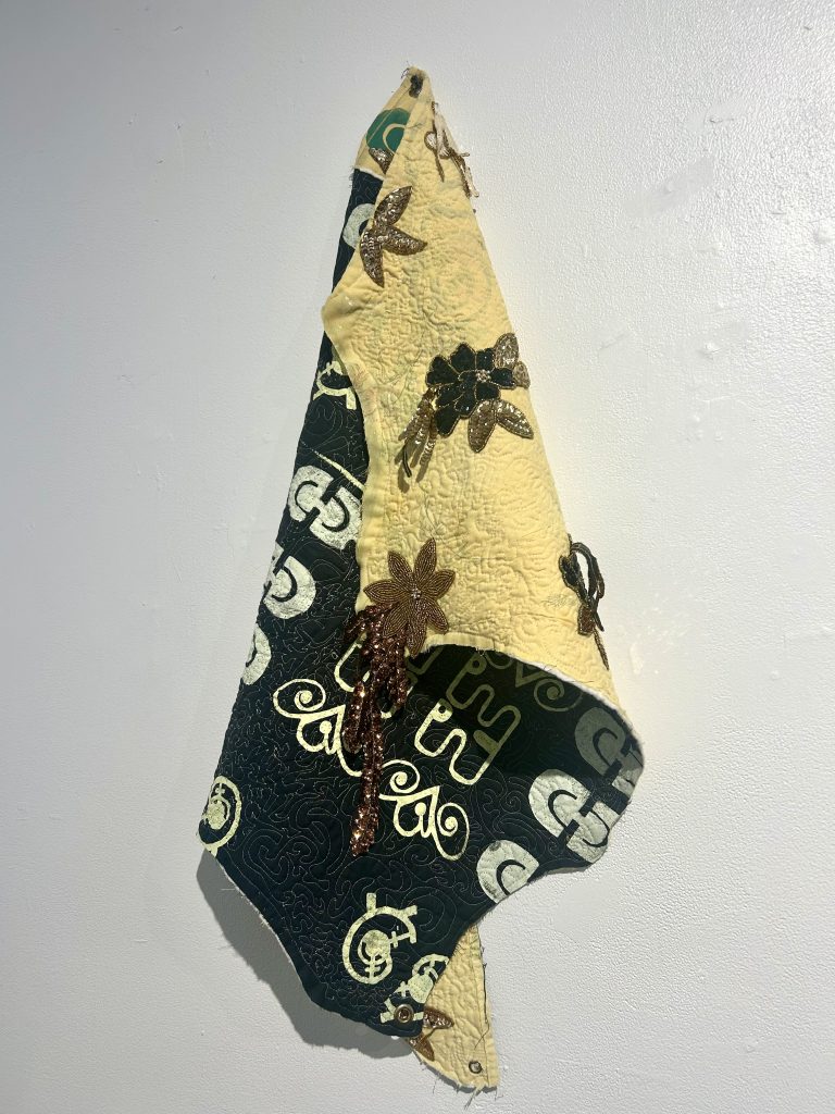 Image: Installation view from We Built This: Making Through Migration. Turn to Your Neighbor, a textile artwork by Mason Brown, hangs on a white wall. The quilt-like piece is draped and hung in a way that shows both sides. The visible yellow side contains flower-like appliqué. The visible black side features African-inspired forms embroidered in yellow and white. Image courtesy of Zhou B Art Center.