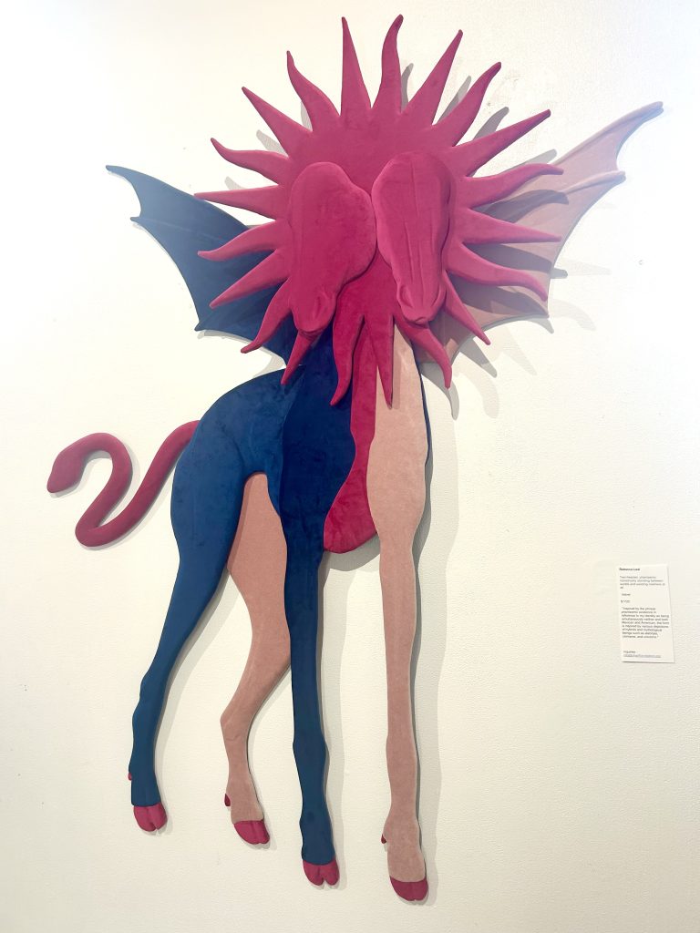 Image: Installation view from We Built This: Making Through Migration. Hung on a white wall is Untitled chimera/monster artwork by Rebecca Leal. The piece is a magenta and blue felted fantasy creature with four legs, two heads, and a tail. A mane of magenta radiates around the heads. Image courtesy of Zhou B Art Center.