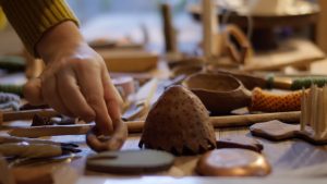 Image: A close up of a tabletop covered in small objects of varying shades of brown. The few objects at the front are in focus while those farther away from the viewer are out of focus. A hand is seen picking up a brown, circular object by pinching it with their thumb and index finger. Image by Ian Carstens.