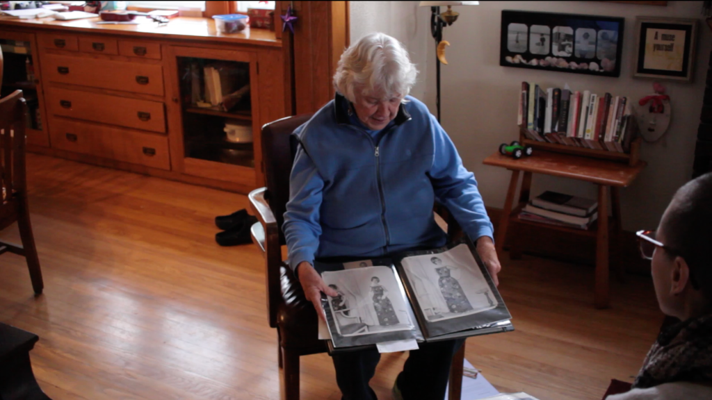 A candid shot of Penny Gardner sitting in a chair in her living room with a large photo album in her lap. The album is open to two images which she shows to Veronica Kirin. Veronica is sitting opposite of her. They both gaze down at the aged, black-and-white images, which depict a woman in a long black gown. Image courtesy of Veronica Kirin.