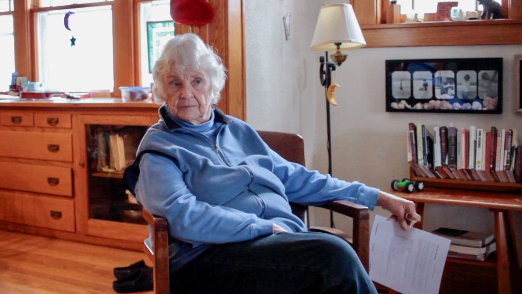 A portrait of Penny Gardner sitting in a wooden chair in your living room. She wears a light blue vest over a light blue sweatshirt. Her legs are crossed and her left arm is resting on the chair's arm. Her face is neutral and tilted slightly left as she looks beyond the camera, to the right of the image. Image courtesy of Veronica Kirin.