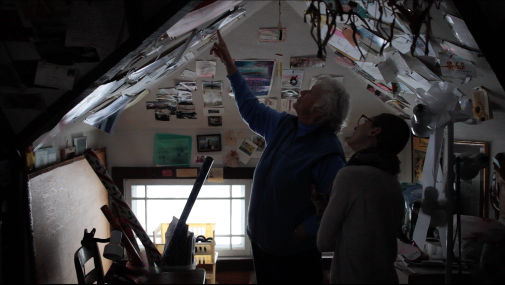 A candid shot of Penny Gardner and Veronica Kirin standing in Penny's home studio/office, framed by a sloped ceiling covered with photos, newspaper clippings, and other memorabilia. They both gaze up as Penny points to a particular image. Image courtesy of Veronica Kirin