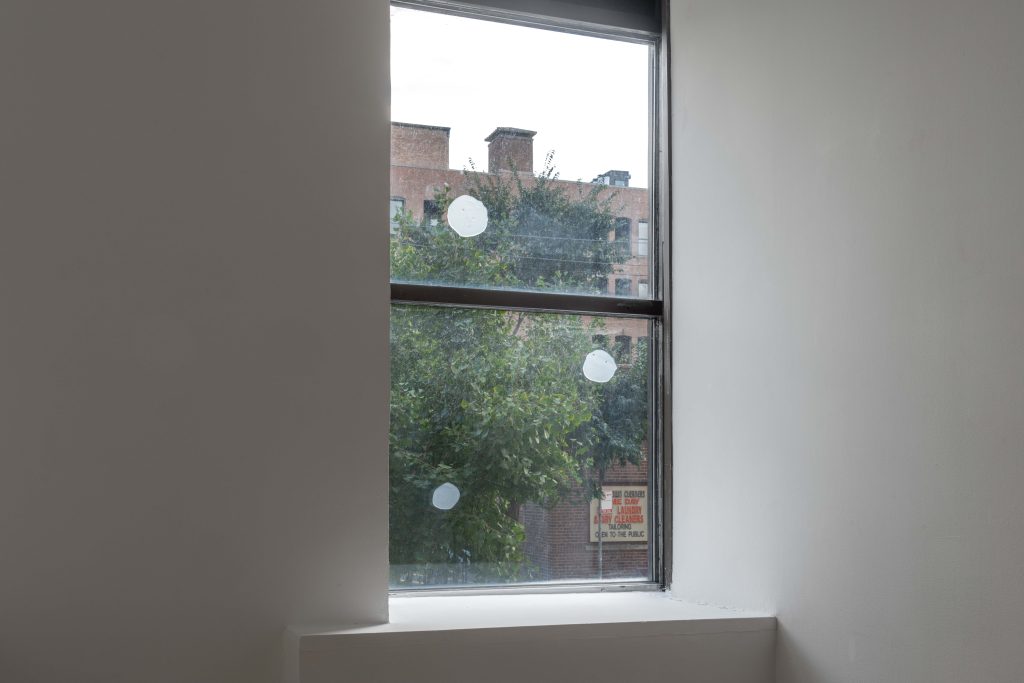 Image: (d)olor atemporal, 2024. Installation view, "A toxin threatens, but it also beckons," Gallery 400. Three translucent circles on the window to a tree, building, and sign outside. Image courtesy of Prairie, Chicago.