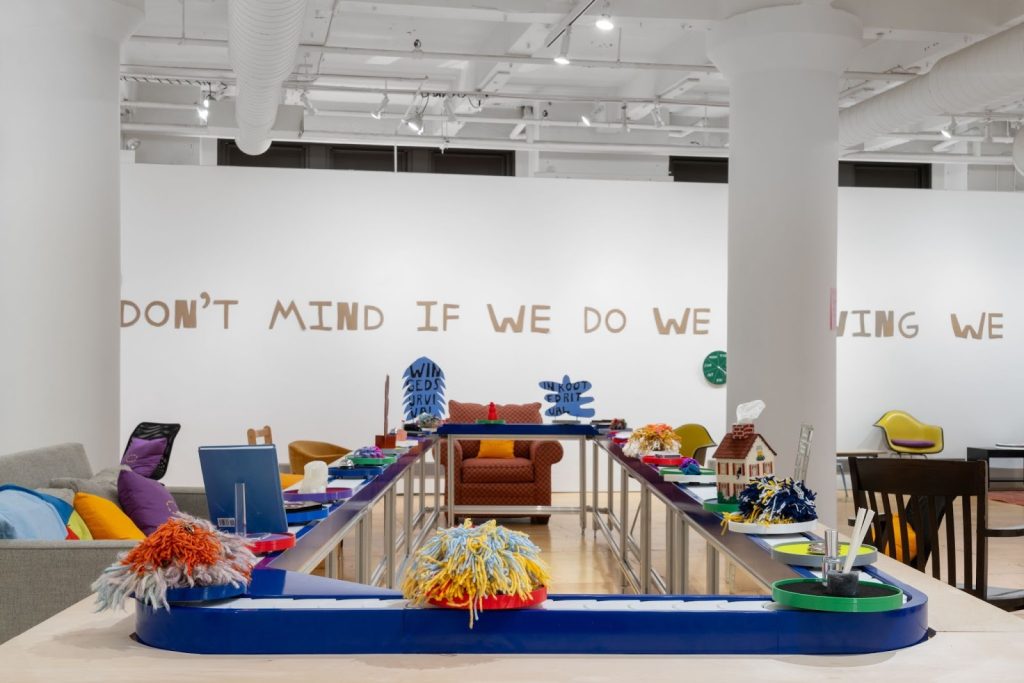 An installation view of Don't Mind if I do, featuring the conveyer belt table centered between a grey couch with colorful pillows and various style chairs placed loosely around the table in the space. The words "don't mind if we do flowing we" hang on the white wall behind the table. Don't mind if I do installation view, 2025. Photo by Jonas Mikowsch Müller-Ahlheim.