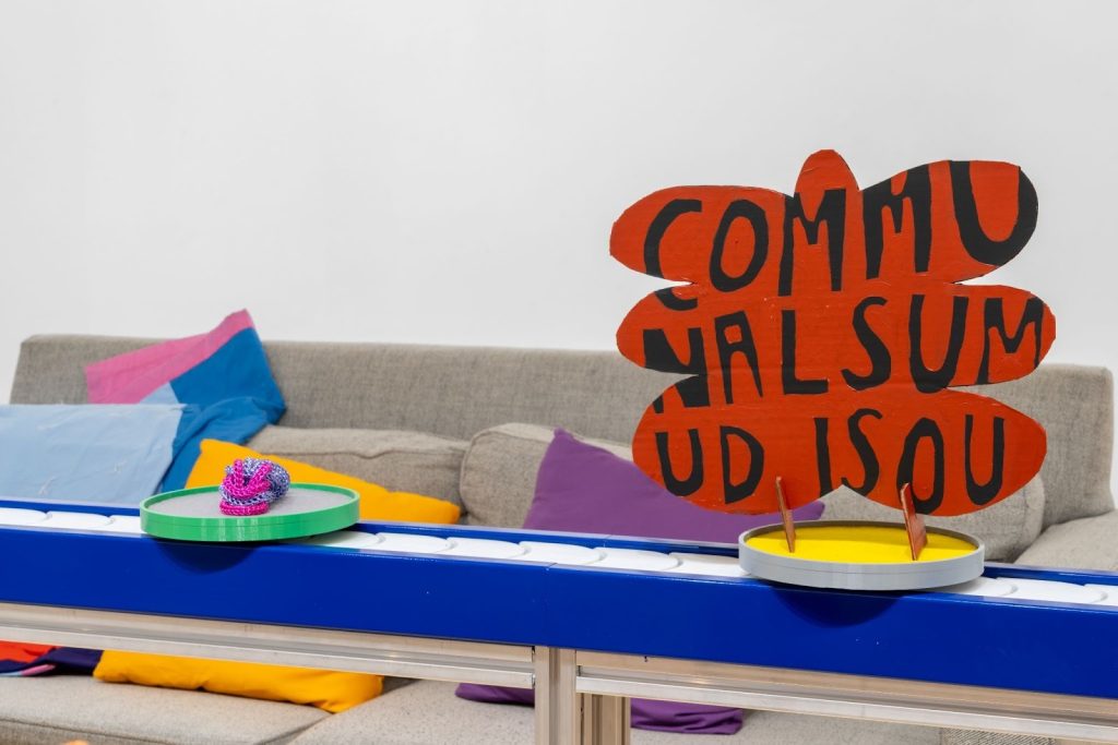 Image: Don't mind if I do installation view, 2025. A closeup of a my access fantasy, which features a red painted cardboard cut out in the shape of a butterfly with the writing "communal sum um isou" in black paint on top of a circular stand. Behind the cardboard cut out is another plate with a soft blue and pink object. Behind the conveyer belt a grey couch with colored fabric and pillows can be seen close by. Photo by Jonas Mikowsch Müller-Ahlheim.