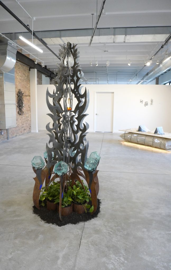 ALTEOKALLI by FÁTIMA can be seen in the foreground, with three pieces by bex ya yolk on view in the background to the right: Protexts I, II, III; stone babies; and The material plane. FÁTIMA's piece IAMHERE III can also be seen in the background on the left. An open gallery space that contains five pieces, including a tall steel sculpture in the foreground, and a steel wall hanging, and a floor-based sculpture. Image courtesy of Chicago Artists Coalition.