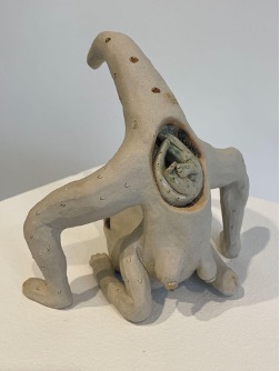 Cecilia Beaven, Cipactil Mamá, 2024, high temperature ceramics, 7”x7”x6”. A gallery podium contains a ceramic sculpture of a surreal reptilian figure with a smaller figure nestled in the stomach. Photo courtesy of the curator Christina Nafziger.