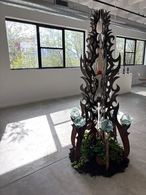 FÁTIMA, ALTEOKALLI, 2025. Earth, water, air, fire, aether, steel, glass, lazurite, selenite, 84”x38”x38”. A steel sculpture standing in the center of the gallery space showered with sunlight from the big gallery windows. Photo Courtesy of Chicago Artists Coalition.