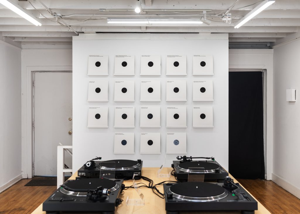 Four turntables sit on top of a wooden table. Nearly twenty records are presented on the gallery wall behind the table.