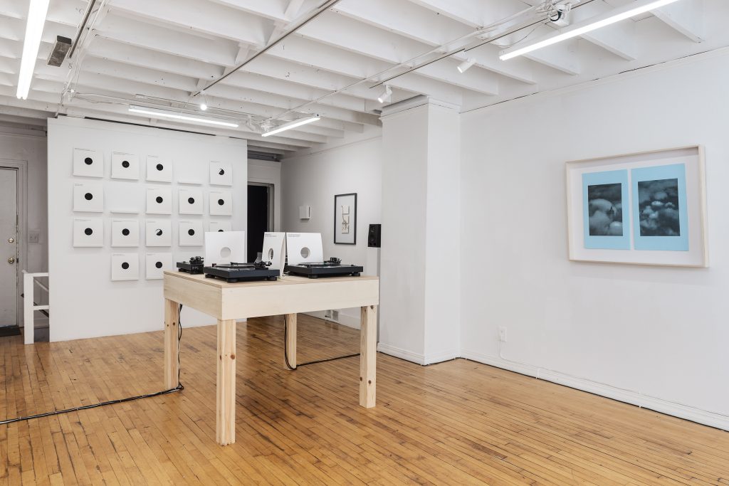 Four turntables sit on top of a wooden table. Nearly twenty records are presented on the gallery wall behind the table. A diptych blue print is hung from the gallery wall on the right side of the image.