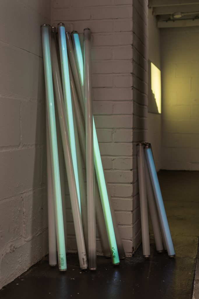 Nearly a dozen fluorescent tubes are stacked upright in the corner of a room and lit by a faint green by an external projector. Four shorter tubes are stacked upright in a doorway.