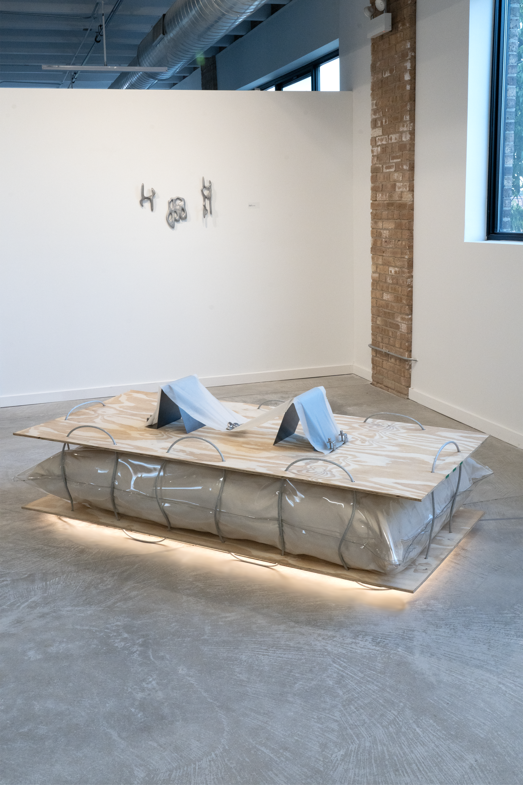 bex ya yolk's piece stone babies sits upon their piece titled The material plane. A floor based sculpture made of plywood, steel wiring, and canvas with an LED light at the bottom separating the piece from the ground. Natural light from the gallery windows spotlights the sculpture. The background features other artworks in the exhibition. Photo courtesy of bex ya yolk.