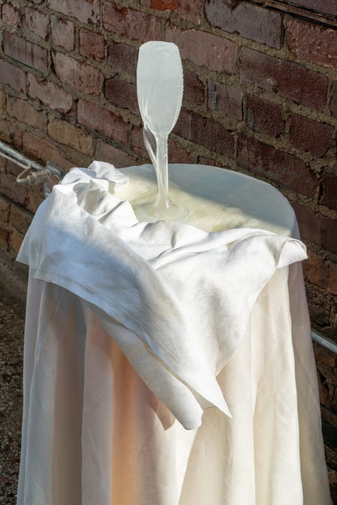Kitty Rauth, As the water rises, (2025). An overflowing champagne glass fountain sits atop a white linen table. The overflowing champagne soaks the linen cloth. Photo by Jonas Mueller-Ahlheim.