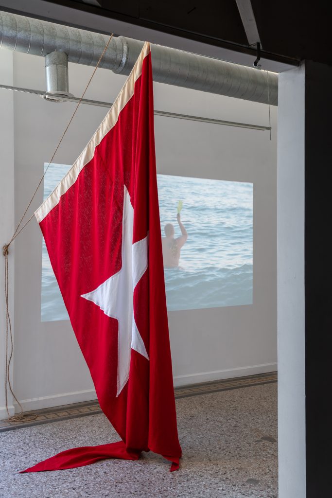 Installation view of "Pleasure Cruise". A large red flag with a white star in the center is suspended from a pillar and the ceiling. Behind the flag there's a video of someone naked and waist-deep in water holding a champagne bottle above their right shoulder. Photo by Jonas Mueller-Ahlheim.