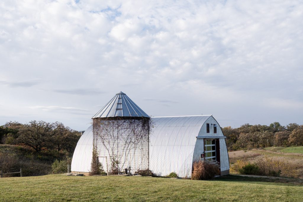 Savannah Sanctuary in Dodgeville, WI. Photograph features a rural landscape. In the center is a silvery, white greenhouse and a cage-like silo. Vines have crawled up the wire frame of the silo and stick out at the top. In the background are small hills covered in grass and trees. The vegetation has a brown dryness to it, indicating fall's arrival. Photo by and courtesy of Max Li.
