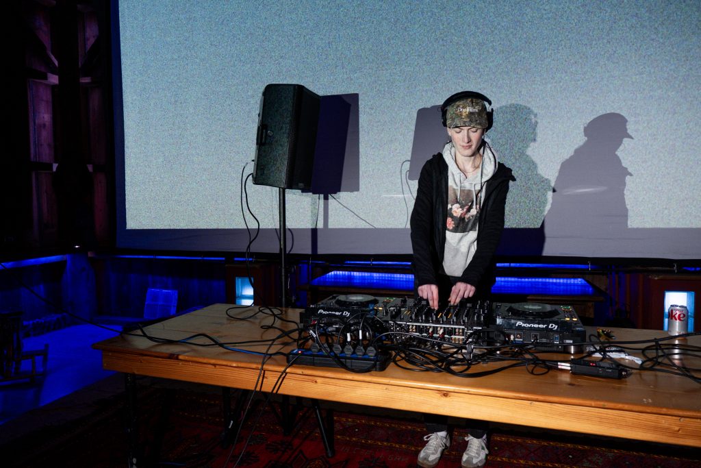 Savannah Sanctuary in Dodgeville, WI. Teddy Sandler DJing on a large wooden table in front of a pixelated projector screen. Blue neon lights illuminate the space. Photo by and courtesy of Max Li.