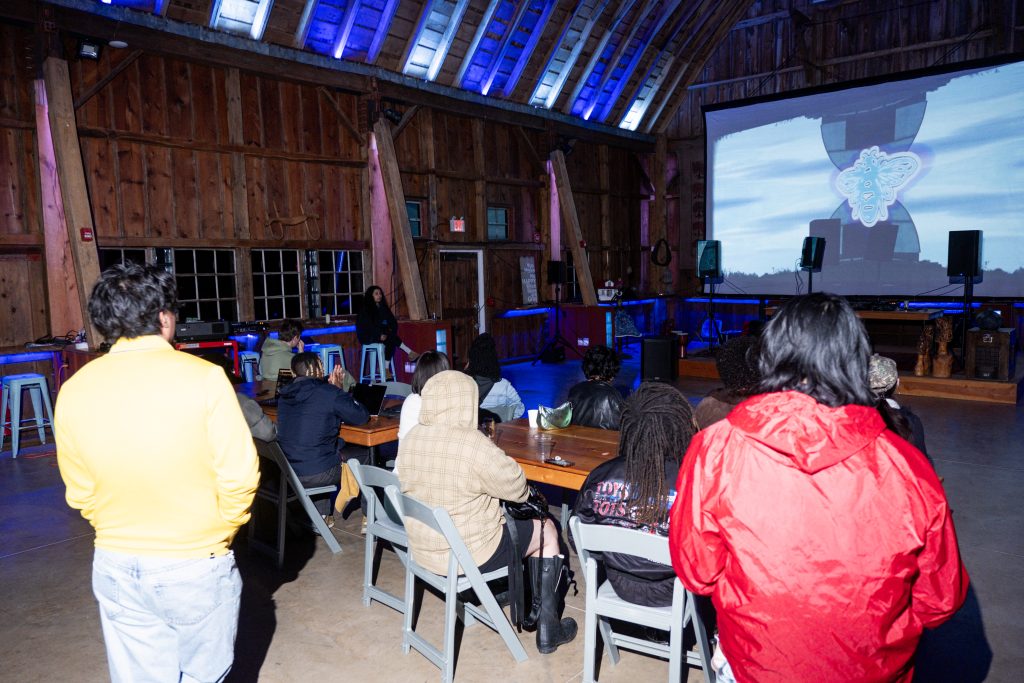 Savannah Sanctuary in Dodgeville, WI. A person is DJing while a projector shines on a large screen. The image from the projector is of a bee with marbled neon colors. The participants watch the performance, some sitting on folding chairs and some standing, their backs are toward the camera. Photo by and courtesy of Max Li.