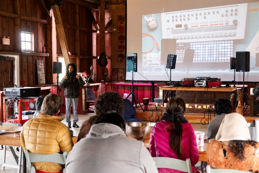 Savannah Sanctuary in Dodgeville, WI. Participants are seated at tables facing the large projector screen, their backs are to the camera. Projected onto the screen are pictures of DJ equipment. A speaker stands at the front, talking to the group. Large black audio speakers, cables, and equipment can be seen around the space,  Photo by and courtesy of Max Li.