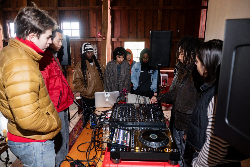 Savannah Sanctuary in Dodgeville, WI. A group of people gather around a wood table covered in DJ equipment. One person is speaking and pointing to the switches while the others listen. Photo by and courtesy of Max Li.