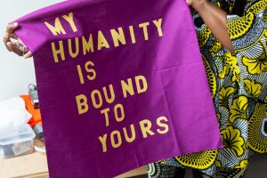 Image: The hands of artist Dorothy Burge can be seen holding a magenta-colored fabric flag with yellow lettering that reads, “My Humanity is Bound To Yours.” Behind the flag you can see glimpses of the bold patterns and bright colors of the outfit Burge is wearing. Photo by Joshua Clay Johnson.