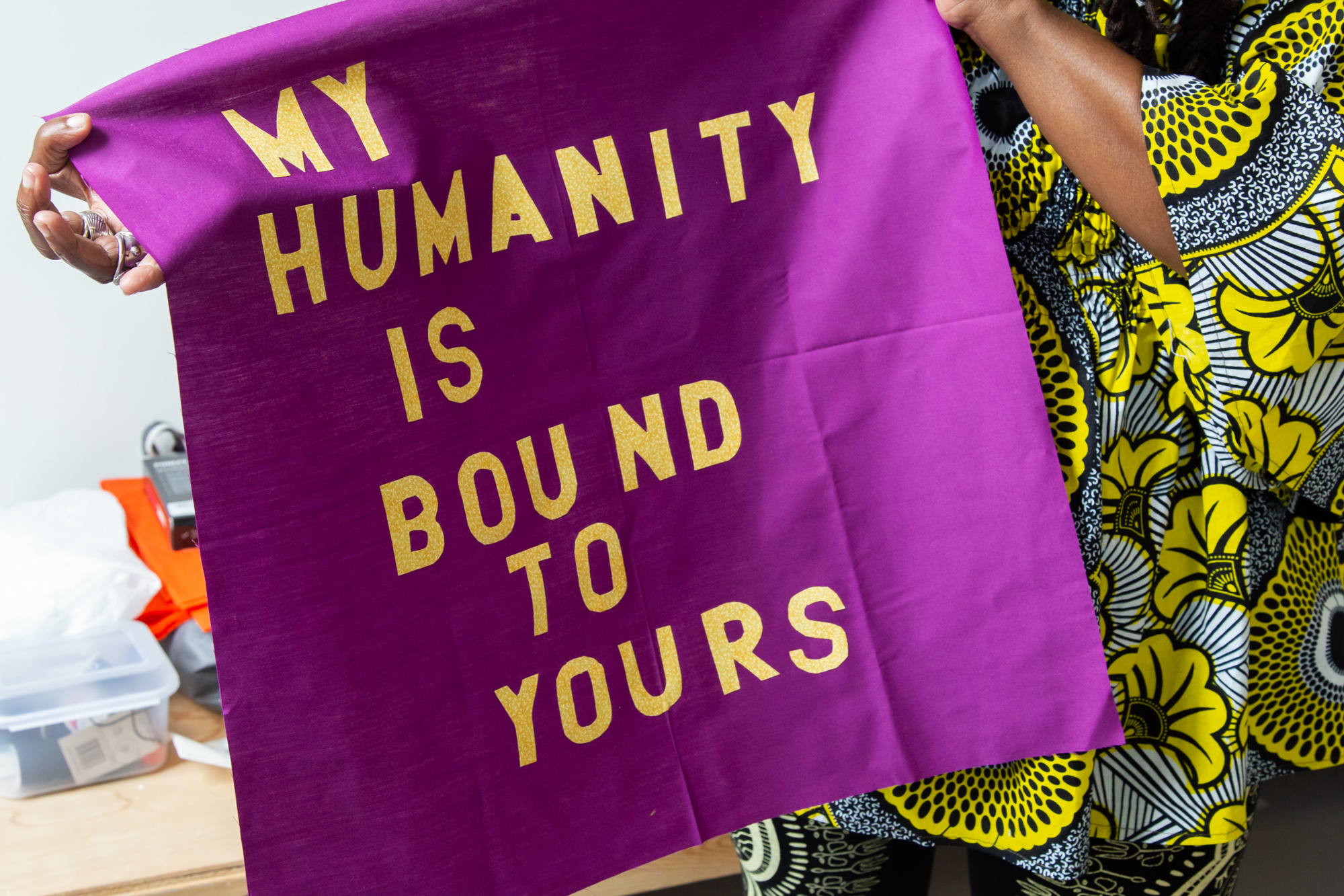 Image: The hands of artist Dorothy Burge can be seen holding a magenta-colored fabric flag with yellow lettering that reads, “My Humanity is Bound To Yours.” Behind the flag you can see glimpses of the bold patterns and bright colors of the outfit Burge is wearing. Photo by Joshua Clay Johnson.