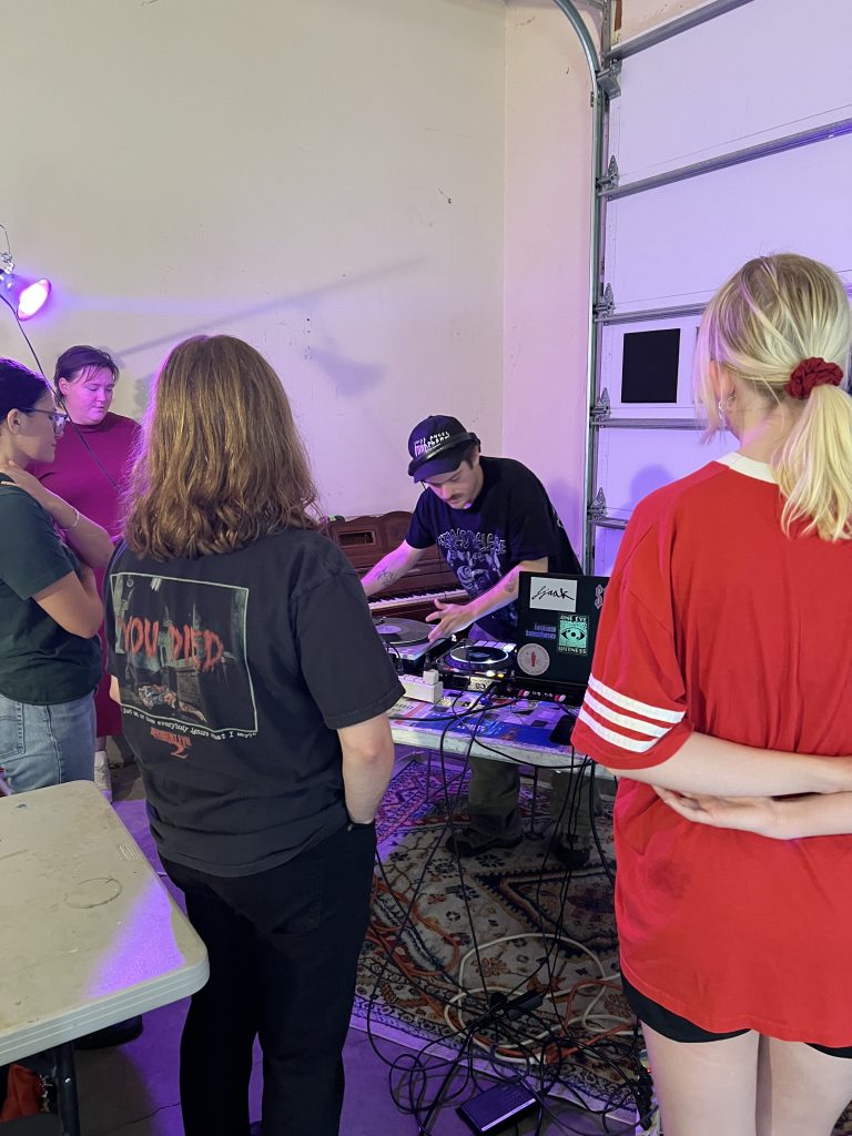 Students gather around a table to observe a workshop led by the DJ duo called Puddle. Tangled cords attached to DJ equipment on the table fall to the floor and rest on a large area rug. A purple light shines overhead. Photo courtesy of Andrés Mora Mata.