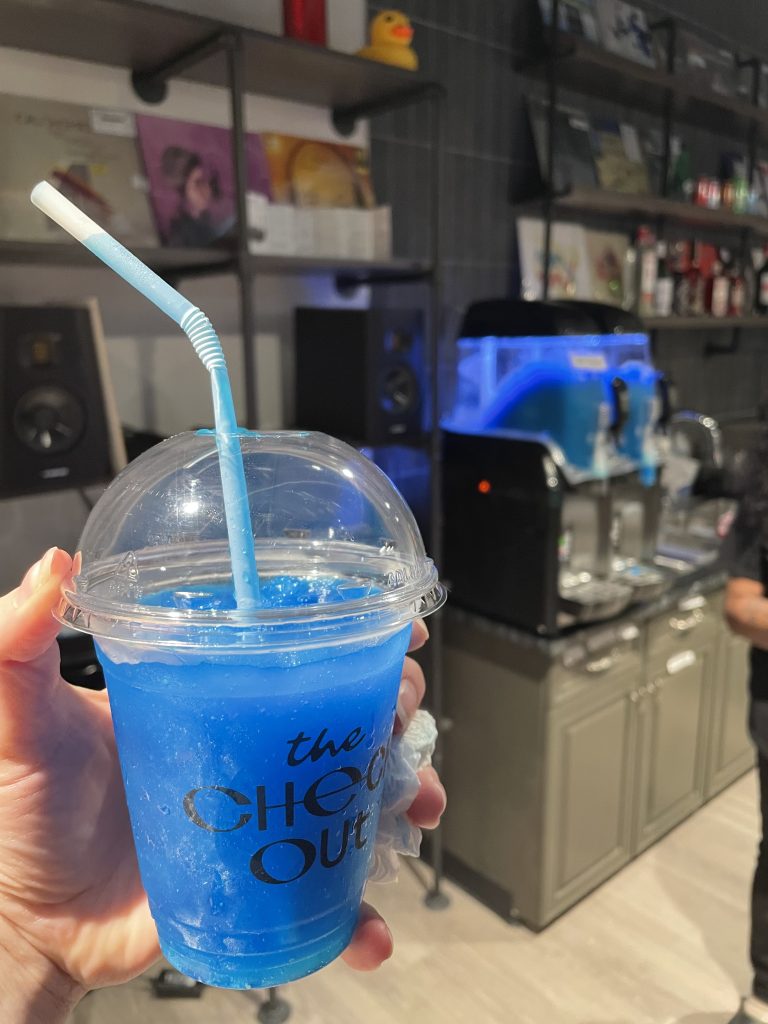 A hand holding a blue slushie with the logo of The CheckOut on the front. In the background are the slushie machines. Image courtesy of author Emily McClanthan. Photo by Alannah Spencer. Courtesy of Access Contemporary Music.