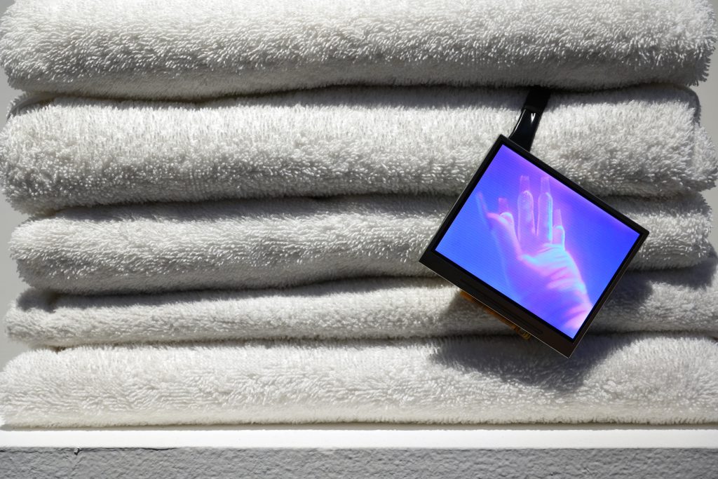 Ále Campos, detail shot of check out time is 4am, (2025). An LED video screen with an image of a gloved hand is presented against a stack of five folded white towels. Photo courtesy of the artist.