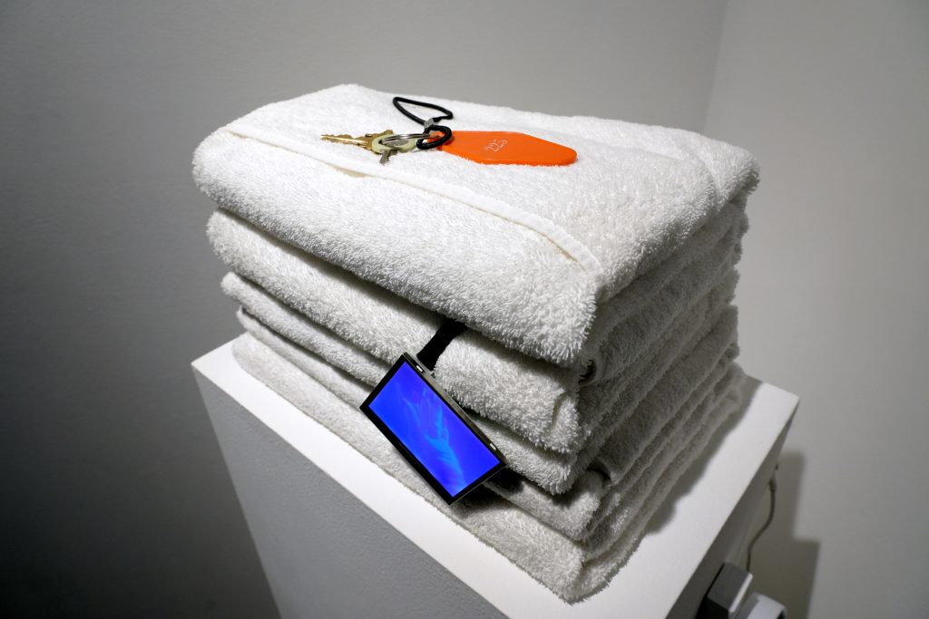 Image: Ále Campos, check out time is 4am, (2025). A stack of five white towels is presented atop a plinth with a locker key and a small LED screen hanging on the side. Photo courtesy of the artist.