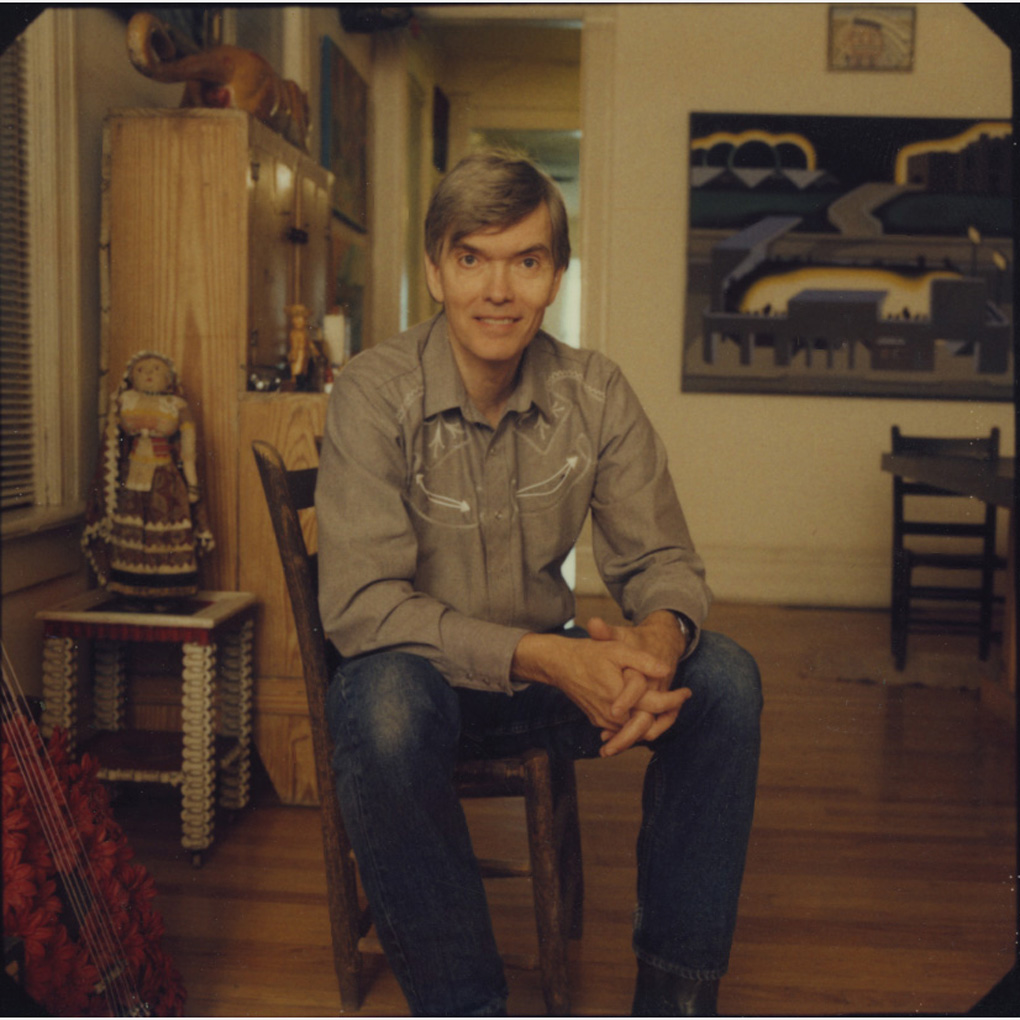 Roger Brown sitting in the second floor living room of his home and studio at 1926 Halsted.The sitter is wearing a gray button down shirt and jeans, he stis leaning forward, hand clasped between his legs, a small smile on his face. The room behind him is decorated with art and wooden furniture.