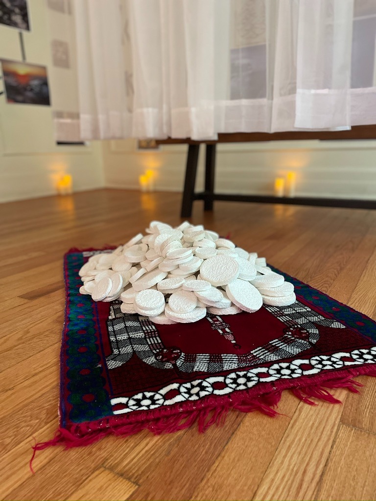 Image: Unclaimed Dua’as, Zeinab Saab, 2019. Textile and mixed media installation. In this work, a prayer rug becomes a site of reflection and reclamation. White circular tokens are placed in a small pile on top of a blue and red prayer rug. The tokens symbolize unspoken or forgotten Islamic prayers, dua’as that hover between memory and invocation. The piece transforms a spiritual tool into a visual object, for shared meditation on faith, identity, and belonging. Photograph by Irina Zadov.