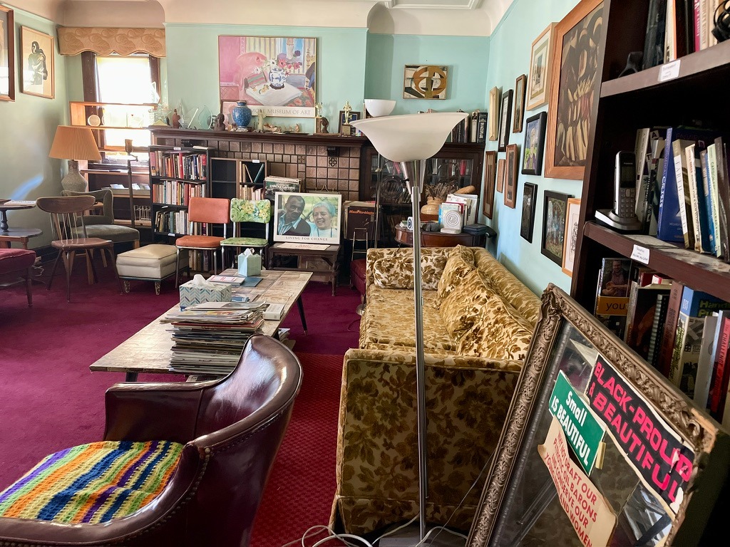 Image: An interior shot of James and Grace Lee Boggs' living room. The walls are teal green, the carpet is maroon, and the room is covered wall-to-wall with books, photos, paintings, and political stickers and buttons. Above the fireplace is a reproduction of a painting from the Detroit Museum of Art. On a chair in front of the fireplace is a portrait of James and Grace Lee Boggs embracing with the words LIVING FOR CHANGE bellow the photo. Photograph by Irina Zadov.