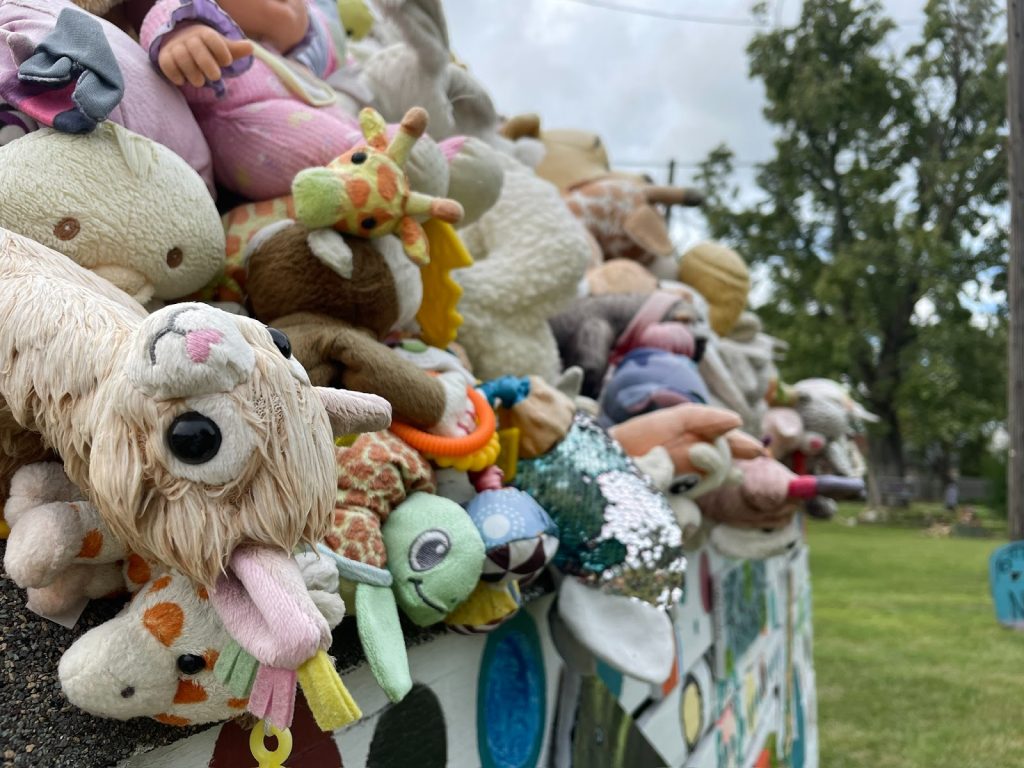 A close up of the Tyree Guyton’s Heidelberg Project featuring a pile of children’s toys soaked by rain and bleached by the sun, attached to the roof of a wooded shack. The toys include a stuffed giraffe, a stuffed turtle, and lots of other stuffed animals covered in pastele fur and silver sequences. In the background there is a green lawn and trees. Photograph by Irina Zadov.