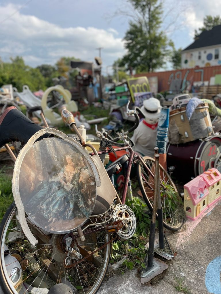 Image: A close up of the Tyree Guyton’s Heidelberg Project featuring a rusted bike, a withered painting of what appears to be a European aristocrat reclining in an armchair holding an open book, with a bent picture frame hanging off it. In the background there are piles of old toys, shoes, books, drums, playground equipment, and a large residential house painted with brightly colored polka dots. Photograph by Irina Zadov