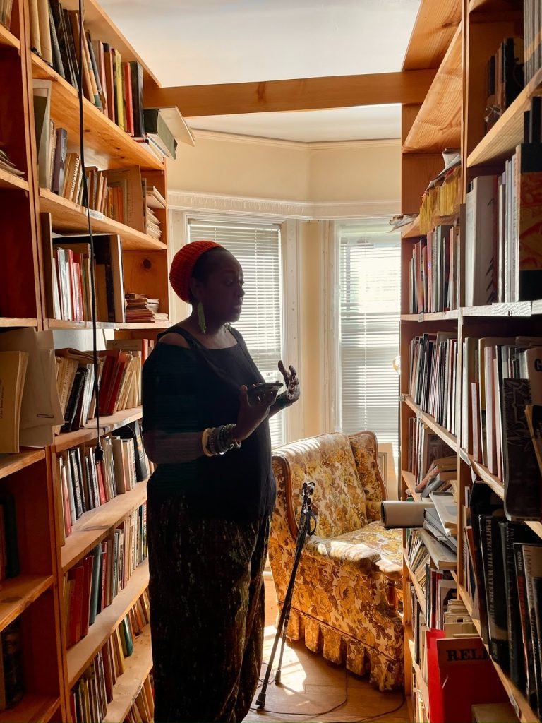 Image: Keisa Davis stands amidst two towering bookshelves overflowing with books of revolutionary history and mass movements across the world. Behind her is a 70s yellow and brown floral print arm chair with Grace Lee Boggs' walking canes leaned up against them. Photograph by Irina Zadov.