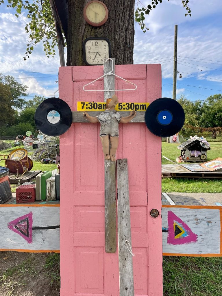 Image: A close up of the Tyree Guyton’s Heidelberg Project featuring a sculpture made out a pink door upon which there are pieces of weather-worn pieces of plywood arranged in a cross with a dilapidated baby doll in the middle which is missing ¾ of its face. On either side of the baby doll there are black plastic records and a yellow sign reading 7:30am - 5:30pm. Above the yellow sign is a white wire hanger. In the background there are several plastic clocks attached to the trunk of a tree, piles of shopping carts, and other discarded objects on a bright green lawn. Beyond that, there’s a bright blue sky with wispy white clouds in the background. Photograph by Irina Zadov.